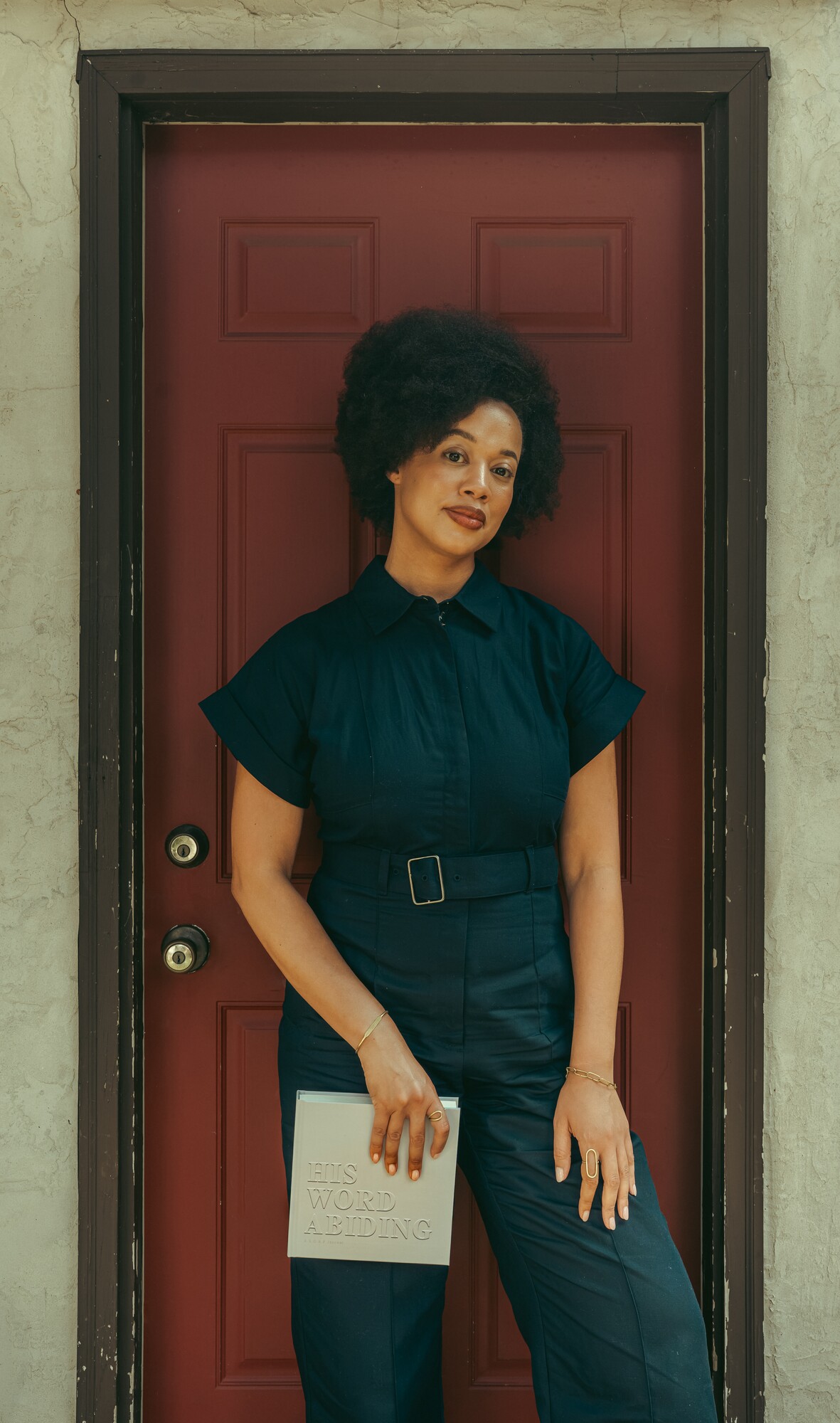 Lauren Ash standing before a deep red door in a belted navy jumpsuit, holding His Word Abiding at her side.