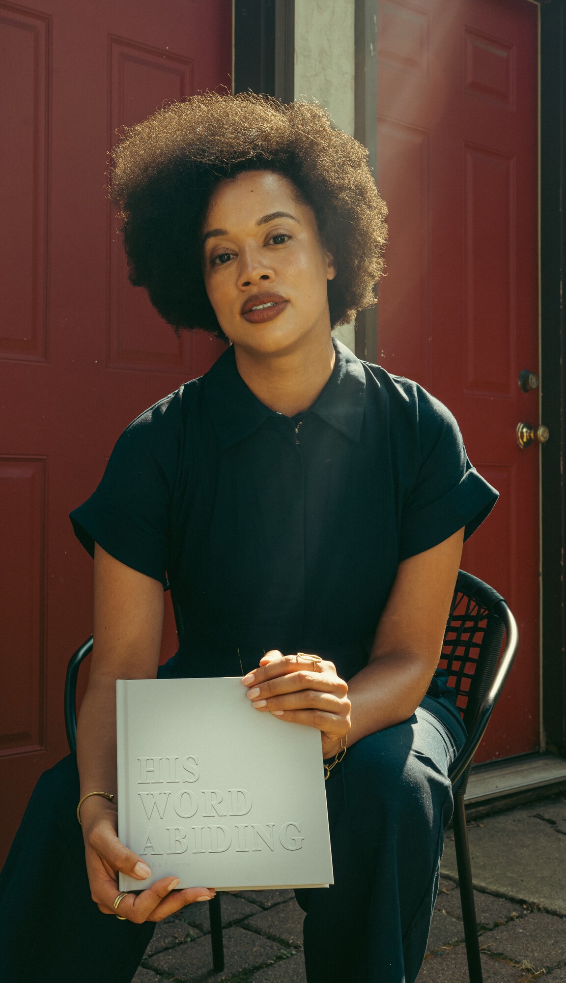 Lauren Ash seated in warm sunlight in front of a red door, holding His Word Abiding in her lap.