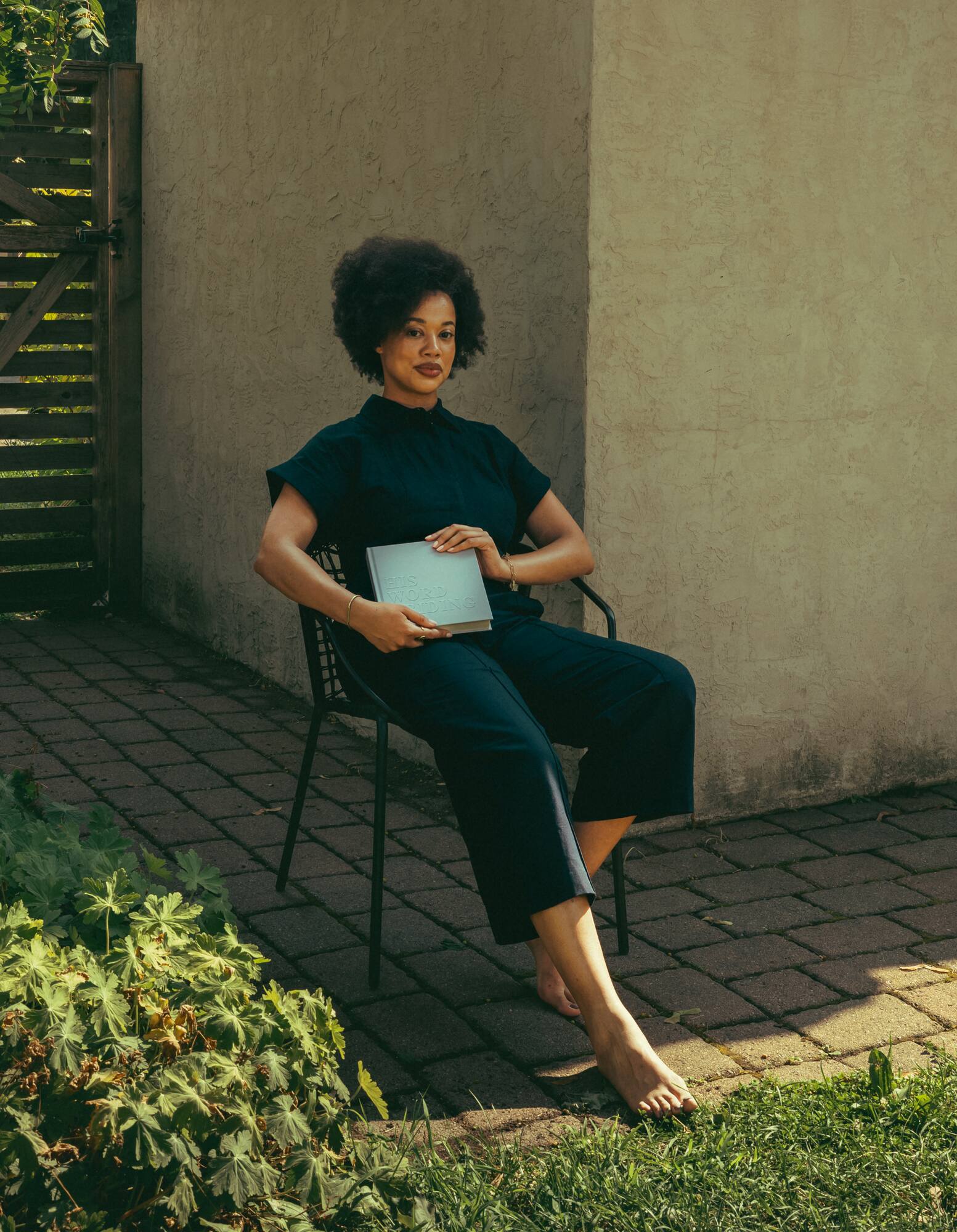 Lauren Ash seated in a courtyard chair in a navy jumpsuit, holding her journal His Word Abiding and looking directly at the camera.